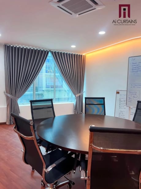A boardroom with an oval table, floor-length drapes drawn across the window and warm light along the ceiling cove.