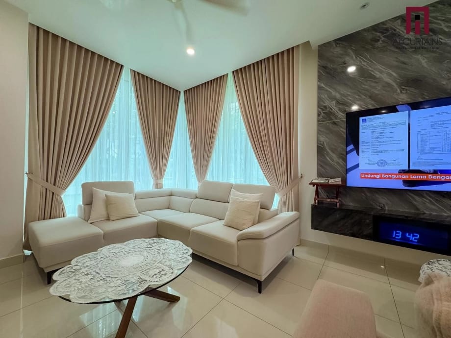 A double-height living room with two tall windows, taupe drapes drawn to the sides on an angle and sheer panels behind them.