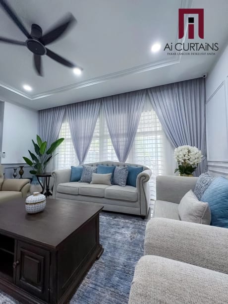 A living room seen lengthways, with sheer panels behind pale blue-grey drapes at the window, two sofas and a ceiling fan.