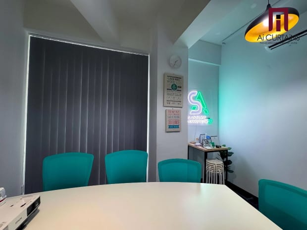A meeting room with grey vertical blinds closed across a full-height window and teal chairs around a pale table.